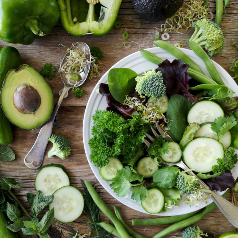 sliced broccoli and cucumber on plate with gray stainless steel fork near green bell pepper, snowpea, and avocado fruit